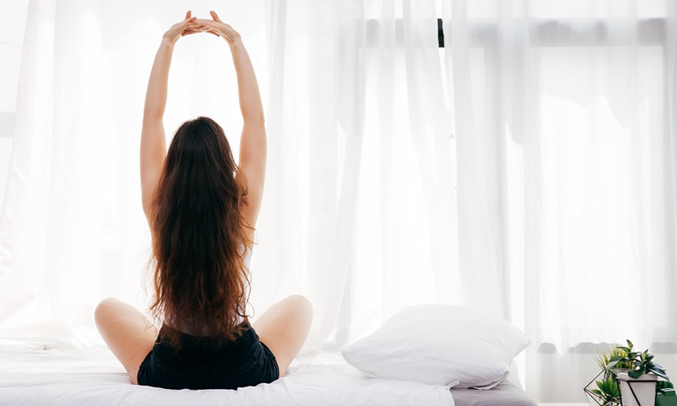 Jeune femme aux longs cheveux bruns de dos qui s’étire assise sur son lit devant une fenêtre